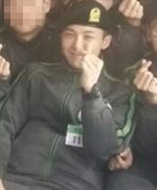 G-Dragon holding up his thumb and index fingers to form a heart, surrounded by fellow soldiers. 