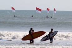 Two surfers on Kuta Beach, Bali carry their boards while Indonesian Military members hoist Indonesian flags riding jet skis in the background, on Tuesday, a day ahead of Independence Day. Thousands of people participated in hoisting 2,112 flags. JP/Zul Trio Anggono