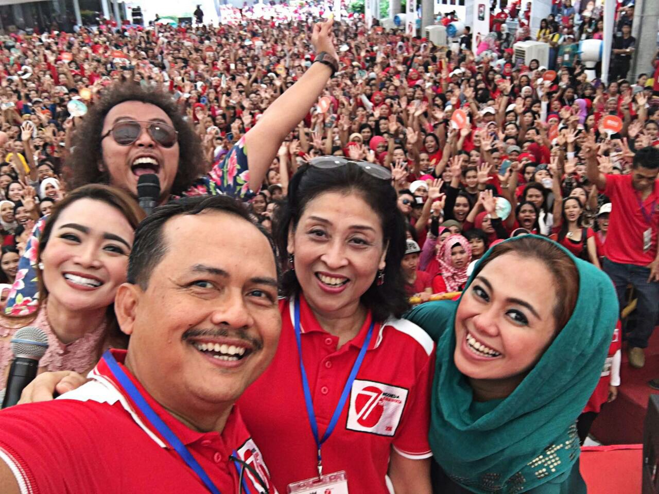  A group of five people are taking a selfie in front of a large crowd. The people in the selfie are all smiling and waving.
