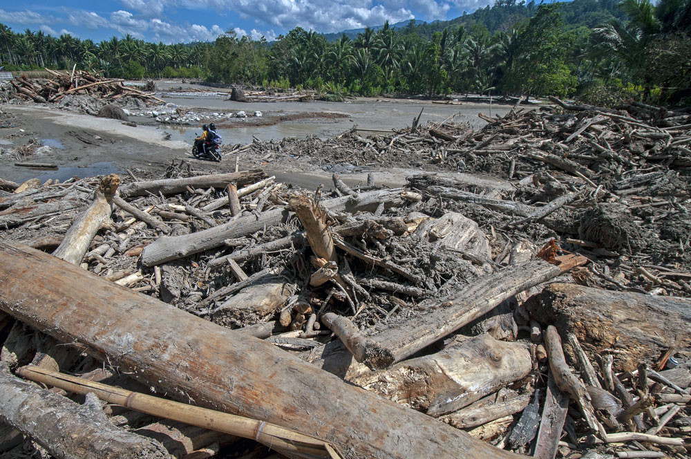 People traveling on a motorcycle use a road flanked by debris left behind by flash floods in Tuva village in Gumbasa district, Sigi, Central Sulawesi, on April 28. Flash floods and landslides frequently occur in the regency.