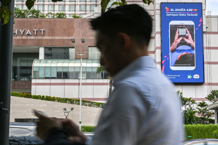 A pedestrian uses his smartphone while walking on a sidewalk in Jakarta on February 28, 2019, past an advertisement for one of Indonesia's top three mobile operators, XL Axiata. 
