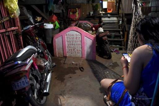 This picture taken on May 21, 2019 shows a woman looking at her smartphone beside a grave inside her home in Phnom Penh. Graveyards may traditionally be the eternal resting place for the dead, but one cemetery in Phnom Penh is increasingly becoming a place to stay for the living as land disputes plague the nation’s poor. 