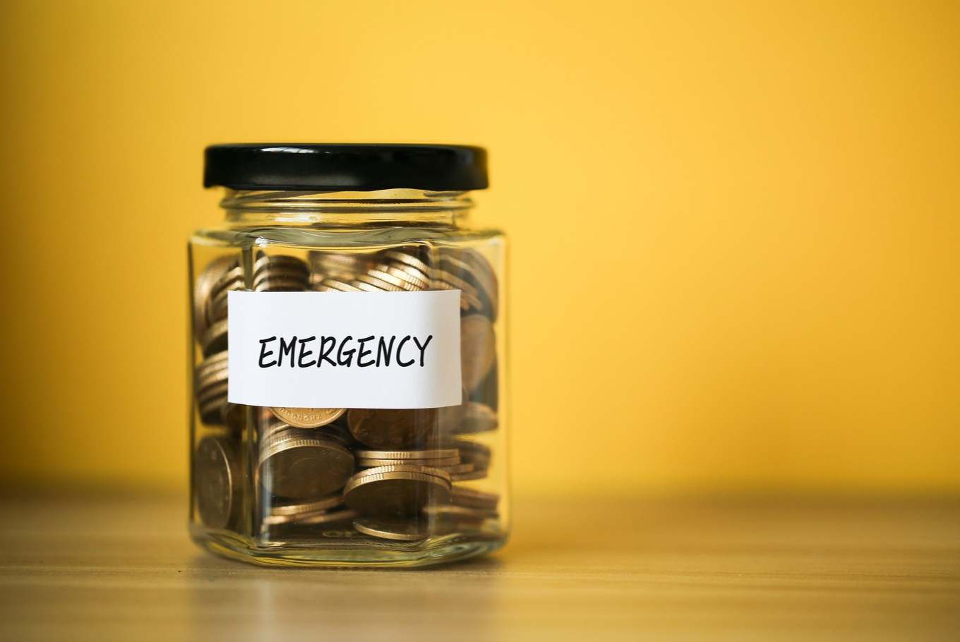  A jar labeled 'Emergency' is filled with coins, sitting on a table against a yellow background.