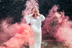 Happy pregnant woman covering her face with smoke grenade outdoors. With the rise of social media, gender reveals have become more and more sophisticated and 'extreme',