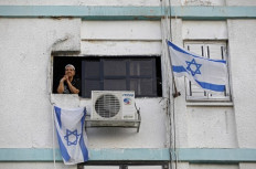 An Israeli woman looks from the window of her home in a building decorated with national flags in the southern city of Ashkelon on April 27, 2020 as the country prepares to mark the Memorial Day for fallen soldiers. - Israel, this evening and until the eve of April 28, will mark the Remembrance day to commemorate the 23,816 fallen soldiers and 3,153 civilians killed in hostilities since 1860, just before the celebrations of the 72th anniversary of its creation according to the Jewish calendar. Official ceremonies will not take place this year due to a national curfew to combat the COVID-19 pandemic. 