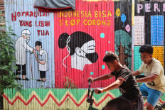 Two boys ride past wall murals promoting public health messages on July 28, 2020 in Kalipasir, Central Jakarta.
