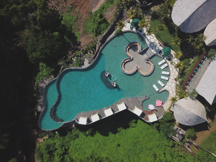 A day by the pool: An aerial view of a split-level swimming pool at Tlaga Singha, a newly opened river country club at Singapadu district in Gianyar regency, Ubud, Bali. The pool mimics the flow of a lagoon cascading into a waterfall to the second level below, where a swim-up pool bar is nestled underneath a rocky grotto.