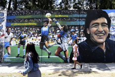 Women walk past a mural by street artist "Uasen" depicting Argentine former football star Diego Maradona -current coach of Gimnasia y Esgrima La Plata football team- scoring the first goal in the FIFA World Cup Mexico 86 match against England, in Villa Palito, San Justo, La Matanza, Buenos Aires province, Argentina, on October 30, 2020 on Maradona's 60th birthday. 