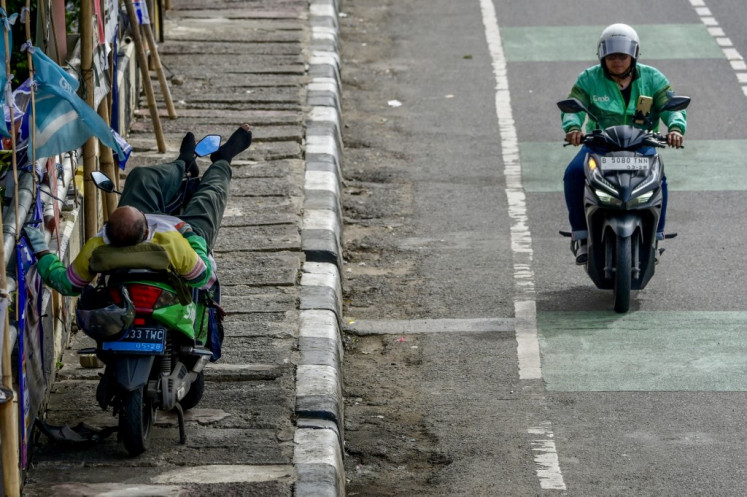 An ojek (motorcycle taxi) driver (left) sleeps on his vehicle beside election campaign posters as a colleague rides past in Jakarta on January 24, 2024, ahead of the country's presidential and legislative polls scheduled to be held on February 14.
