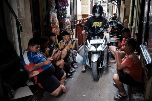 A motorcyclist rides through an alley in Jakarta on Jan. 21, 2024, past a group of boys playing online games on their smartphones.