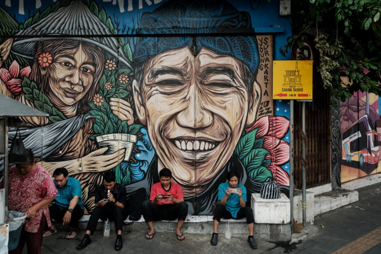 People use their mobile phones as they sit in front of a graffiti depicting an image of President Joko “Jokowi“ Widodo on a street in Surakarta, Central Java, on July 31, 2024.