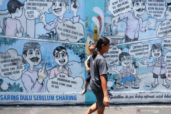 A woman carries a toddler on April 6, 2024, as she walks past an anti-hoax and anti-hate mural on a street in Hepi kampung of Joho subdistrict in Surakarta, Central Java.

