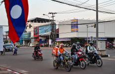 People drive their motorbike near a flag of Laos at the entrance of the National Convention Center in Vientiane on October 7, 2024, ahead of the 44th and 45th ASEAN Summits.