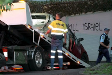 This frame grab taken from video footage provided by the Australian Broadcast Corporation (ABC) on December 11, 2024 shows a police officer walking past a torched car being removed in front of anti-Israel graffiti written on a wall in the Sydney suburb of Woollahra. 
