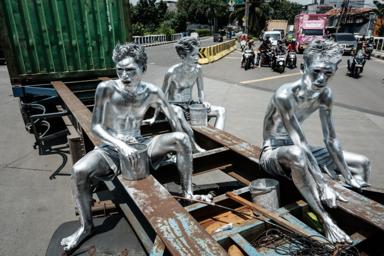 This photo taken on April 7, 2025 shows street performers Keris Munandar (right), his brother Ari Munandar (left), and their neighbor Riyan Ahmad Fazriyansah hitch a ride on the back of a container truck to reach an intersection in Jakarta. On a rainy day in Indonesia's capital Jakarta, three men coated in metallic paint known as the “manusia silver“, or silvermen, brave the elements at an intersection near a mall to ask drivers for change.
