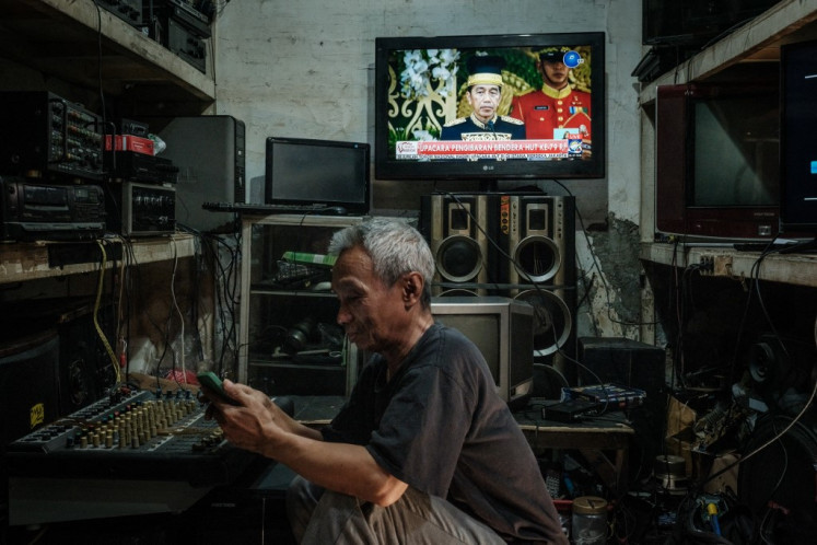 A man uses a smartphone while a television screen shows a live broadcast of Indonesia's 79th Independence Day celebrations from the future presidential palace in the future capital Nusantara (IKN), at his electrical recycling workshop in Jakarta on August 17, 2024. 