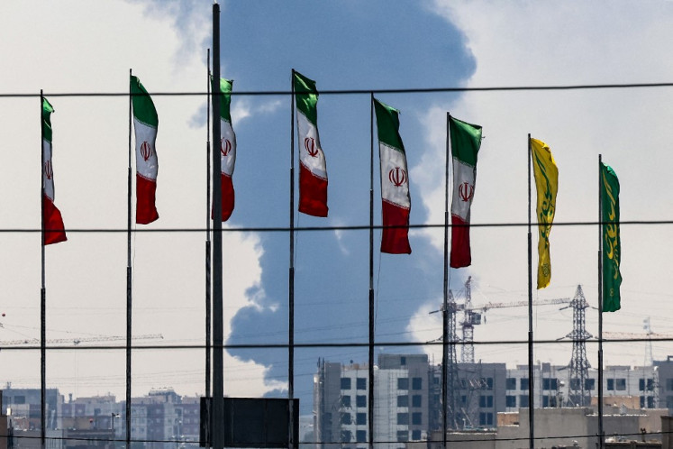 Flags flutter along a bridge as a plume of heavy smoke and fire rise from an oil refinery in southern Tehran after it was hit in an overnight Israeli strike on June 15, 2025. Iran fired a fresh barrage of missiles at Israel, state television announced early on June 15, as the rivals exchanged fire for a third day.
