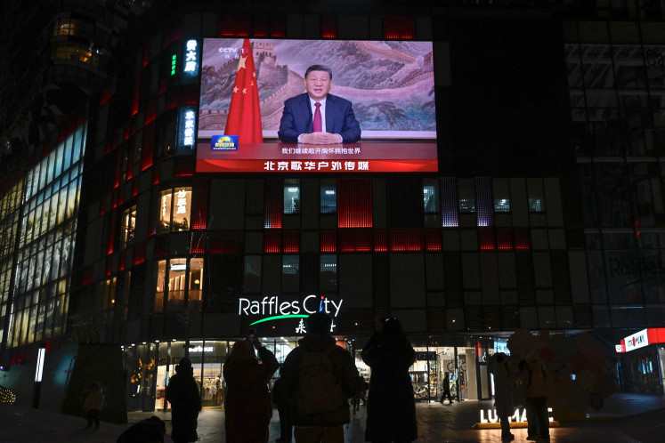 People stand beneath a large screen showing China's President Xi Jinping delivering a speech ahead of the New Year's Eve celebrations in Beijing on Dec. 31, 2025.