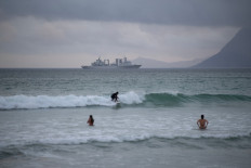 A surfer rides a wave with the Chinese comprehensive supply ship Taihu (Hull 889) in the background in False Bay, close to Simon's Town, near Cape Town, on January 6, 2026. 