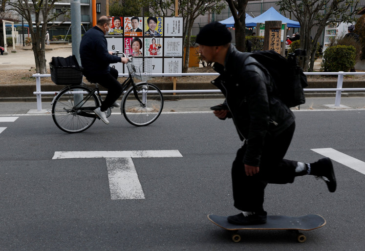 A man on a skateboard crosses a street as a cyclist rides past election campaign posters for the Feb. 8 snap election, displayed on a board in Tokyo on Feb. 6, 2026.