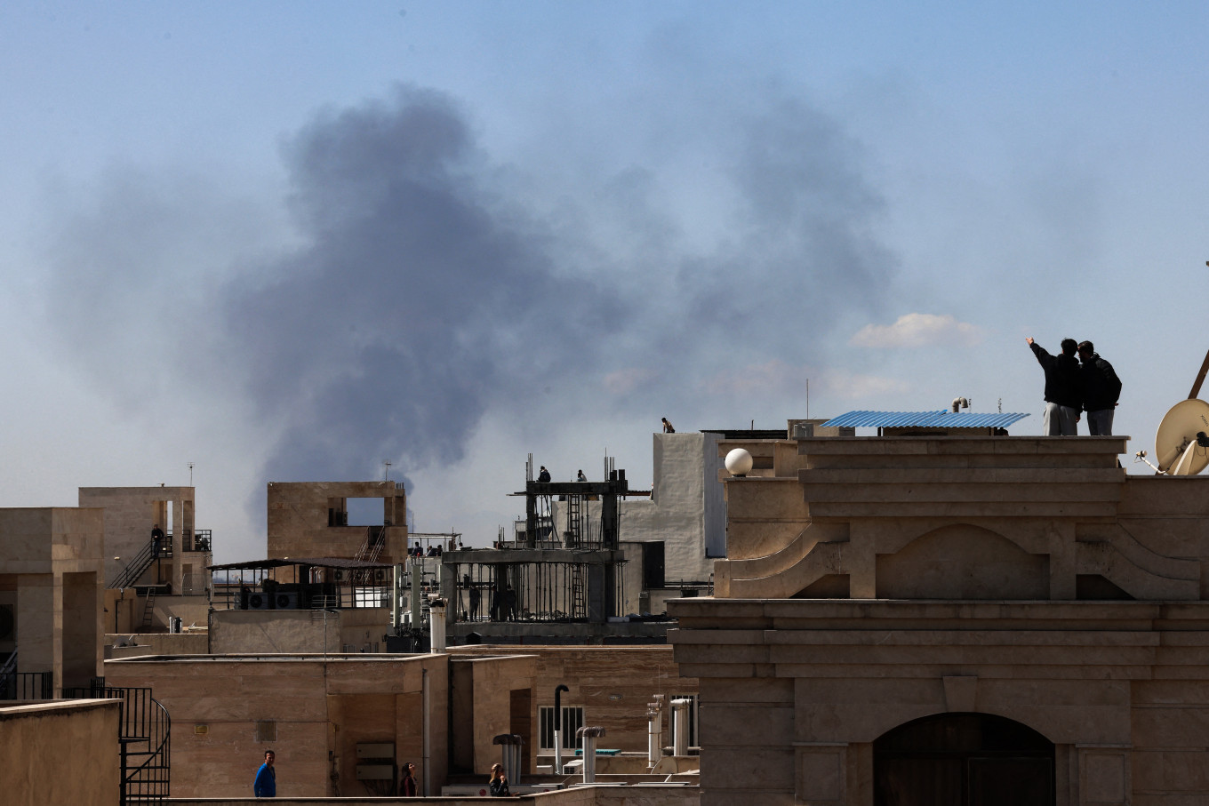 People stand on the roofs of their houses in Tehran and watch plumes of smoke rise from a series of explosions on March 1, 2026, following the opening salvo of the joint United States-Israeli attacks on Iran.