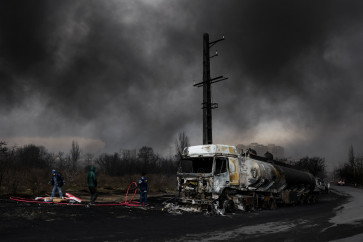 People stand near a destroyed vehicle on March 8, 2026, as smoke rises after a reported strike on the Shahran fuel tanks in Tehran, amid the United States-Israel war on Iran.