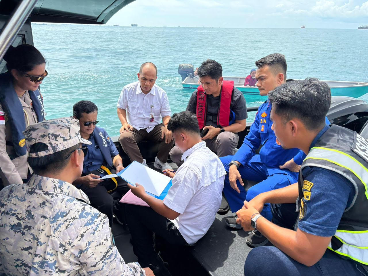 A diplomat from the Indonesian Consulate General in Johor Bahru, Malaysia, signs a handover letter while surrounded by officials from various Indonesian agencies and observed by a Malaysian Maritime Enforcement Agency (MMEA) officer (left, sitting) during a rendezvous on May 7 on the sea border between the countries. The consulate general repatriated two fishermen and their fishing boat (background) who drifted away into Malaysian waters after their boat ran out of fuel.