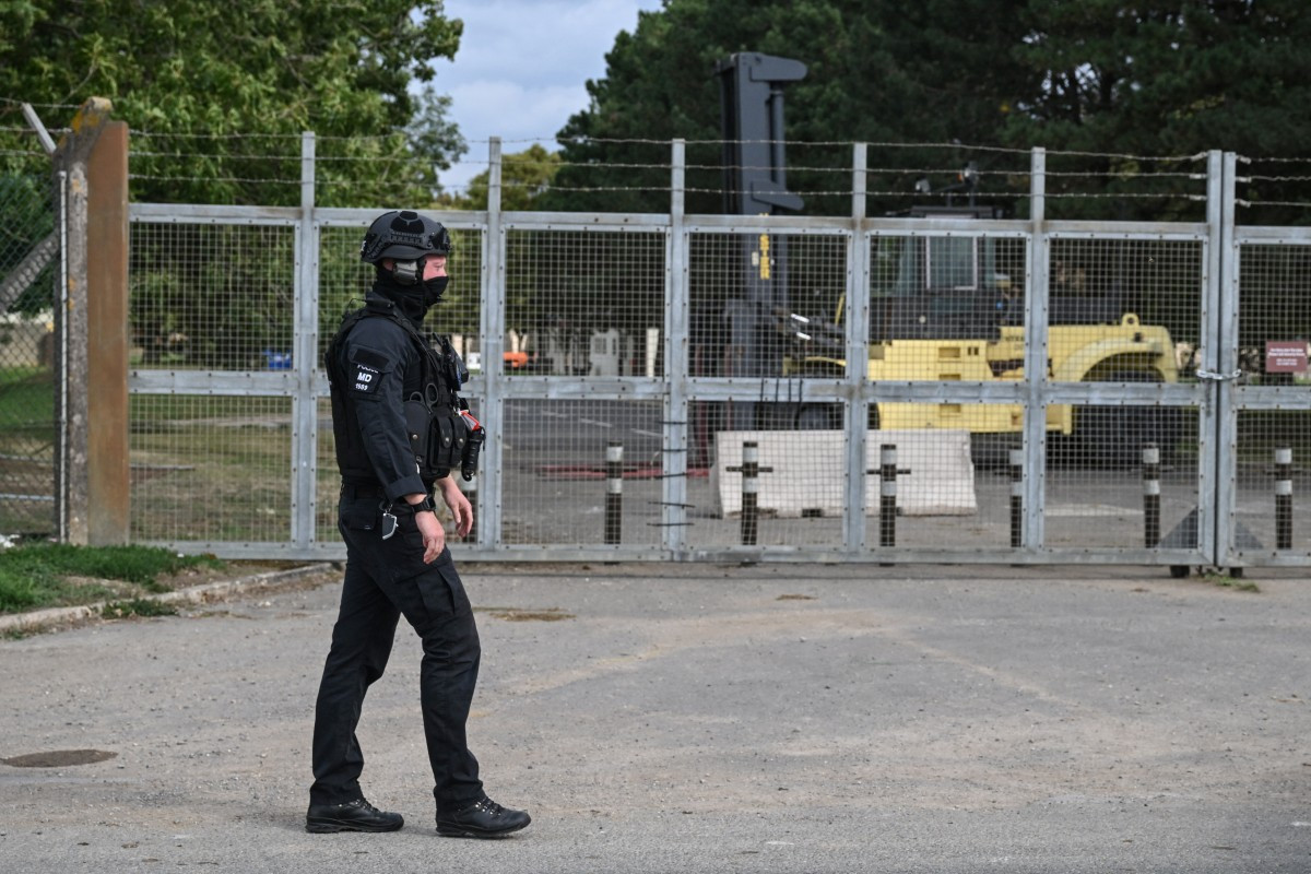 A masked policeman guards an entrance to RAF Fairford base, blocked by heavy machinery (back right) inside the gate, in Fairford, south-west England on September 27, 2026, as UK counterterrorism police investigate an incident near the base in the village of Whelford. British police on September 27 declared a major incident and evacuated villagers living near a base used by the US air force after arresting several men on suspicion of explosives offenses.