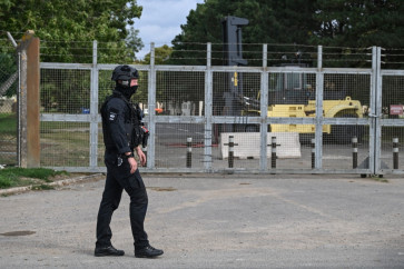 A masked policeman guards an entrance to RAF Fairford base, blocked by heavy machinery (back right) inside the gate, in Fairford, south-west England on September 27, 2026, as UK counterterrorism police investigate an incident near the base in the village of Whelford. British police on September 27 declared a major incident and evacuated villagers living near a base used by the US air force after arresting several men on suspicion of explosives offenses.